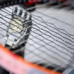 Zbliżenie na rakietę tenisową podczas wymiany naciągu. Widać metalowe przelotki i fragmenty naciągu.