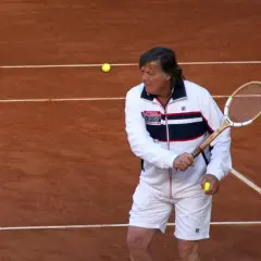 Panatta w białej kurtce i spodenkach, z rakietą i piłką tenisową na korcie.