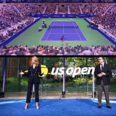 Dwie osoby stoją na niebieskim dywanie przed ekranem wyświetlającym mecz tenisowy US Open.