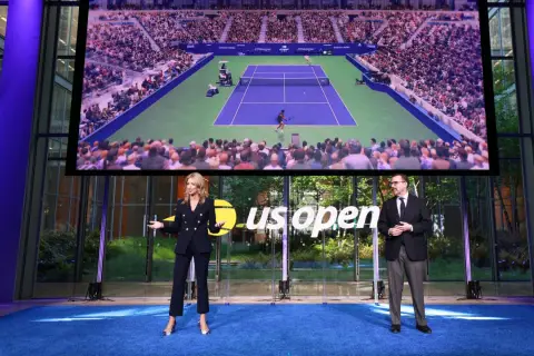 Dwie osoby stoją na niebieskim dywanie przed ekranem wyświetlającym mecz tenisowy US Open.