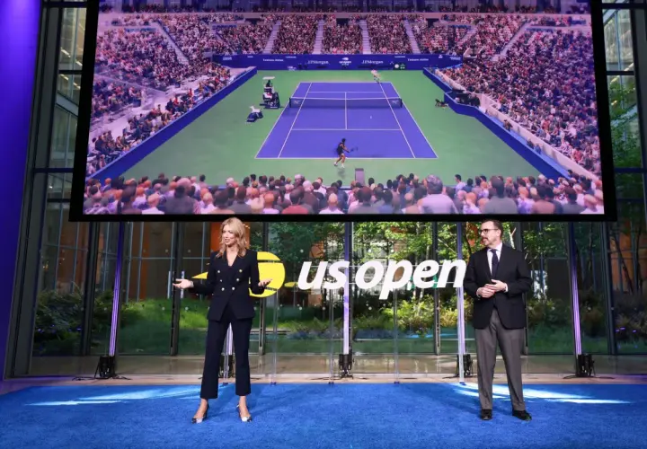Dwie osoby stoją na niebieskim dywanie przed ekranem wyświetlającym mecz tenisowy US Open.
