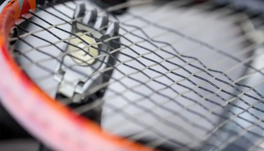 Zbliżenie na rakietę tenisową podczas wymiany naciągu. Widać metalowe przelotki i fragmenty naciągu.