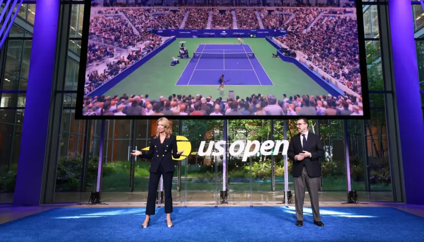Dwie osoby stoją na niebieskim dywanie przed ekranem wyświetlającym mecz tenisowy US Open.