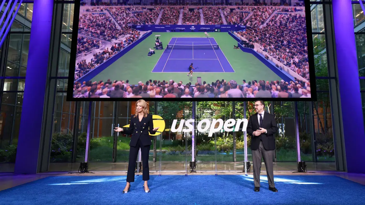 Dwie osoby stoją na niebieskim dywanie przed ekranem wyświetlającym mecz tenisowy US Open.