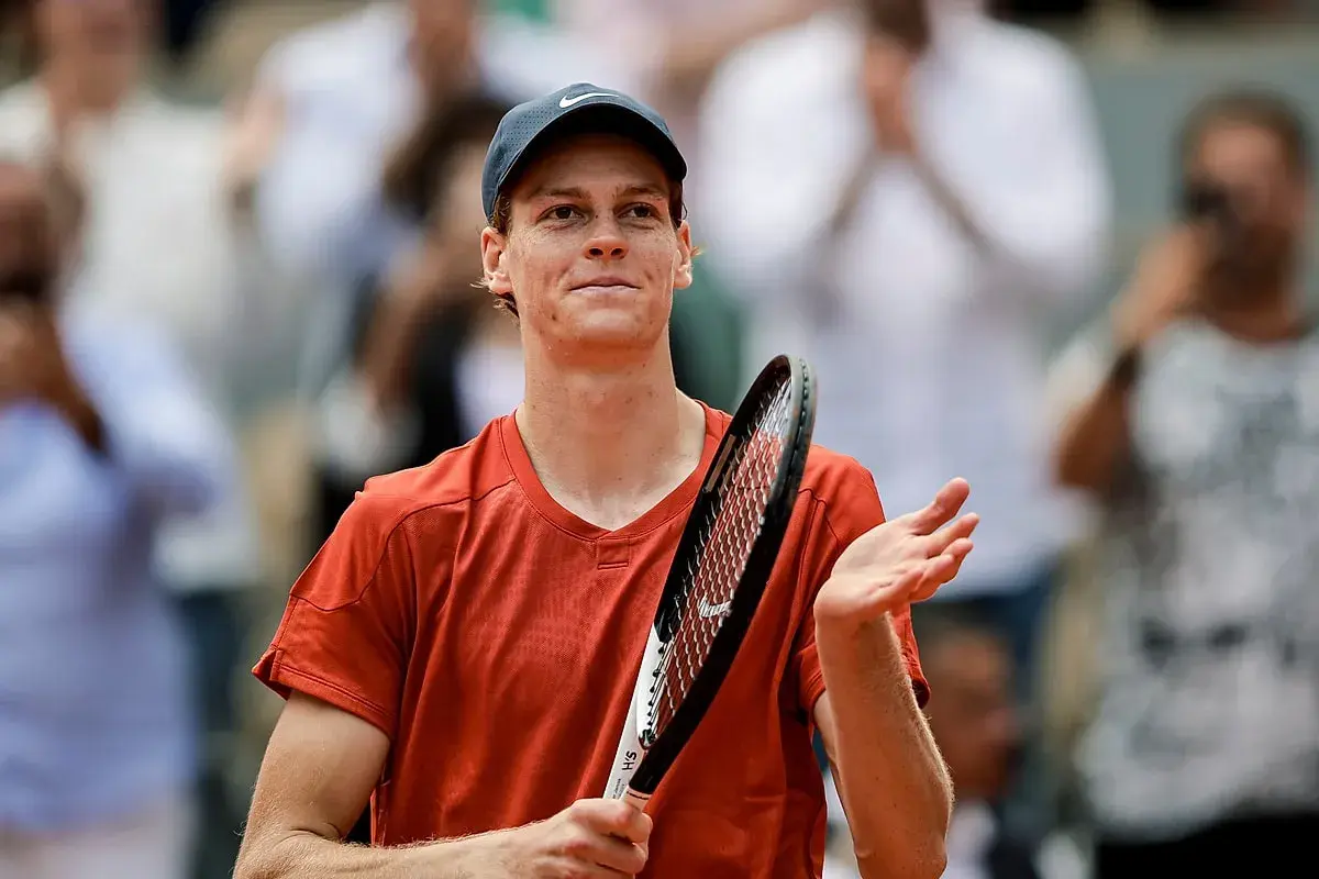 Tenisista w pomarańczowej koszulce i czapce z daszkiem, z rakietą w dłoni, dziękuje publiczności po meczu ATP 250.